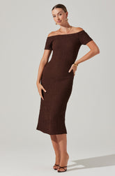 A woman is wearing a fitted, off-the-shoulder, short-sleeved, midi-length dress in a rich, dark brown color. The dress has a textured, woven pattern throughout. She is accessorized with large hoop earrings, a delicate necklace, a bracelet, and rings. She is also wearing strappy heeled sandals.